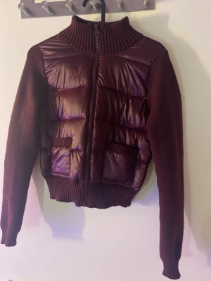 Vinröd cardiganjacka med quiltad front - Mysig vinröd cardiganjacka med stickade ärmar och rygg samt quiltad, glansig front. Jackan har hög krage, dragkedja och två fickor framtill. Perfekt för lager-på-lager och ger en cool, sportig look. Materialmix av stickat och vadderat tyg.