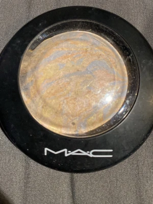 MAC  shade lightscapade  - Säljer en MAC mineralize bronzer med marmorerad yta i nyanser av guld, beige och ljusblått. Kommer i en rund svart dosa med genomskinligt lock och MAC-logga. Perfekt för att ge ansiktet en naturlig glow och värme.