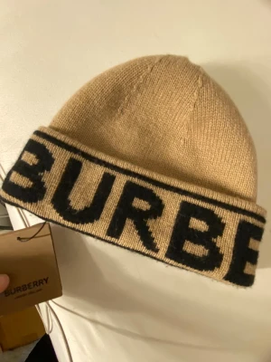 Beige mössa från Burberry - Snygg beige stickad mössa från Burberry med bred uppvikt kant och stor svart logotyp runt hela kanten. Tillverkad i mjukt material som ger en lyxig känsla och passar perfekt till höst och vinter. Klassisk och trendig accessoar för dig som gillar designerplagg. Den kommer tyvärr inte till användning den är äkta det finns kvitto köpte den på NK. Köpte den för 3000kr och säljer den för 2000 eftersom den är ny och äkta. Pris kan diskuteras! 