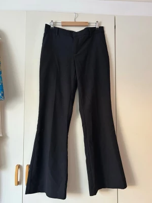 Svarta utsvängda kostymbyxor - Säljer ett par svarta kostymbyxor med utsvängda ben och hög midja. Byxorna har klassisk design med fickor fram och bälteshällor. Perfekta för en stilren och trendig look. Materialet känns mjukt och följsamt.