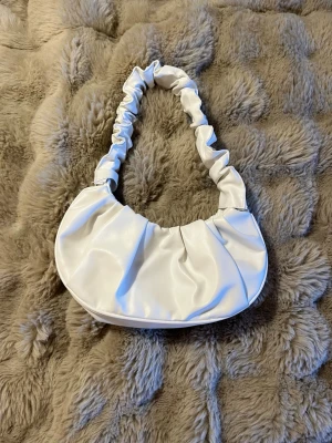 Vit scrunchie-handväska i fejkläder - Trendig vit handväska i fejkläder med scrunchie-inspirerat handtag och mjuk, rundad form. Väskan har svart innerfoder och stängs med magnetknapp. Perfekt accessoar för att lyfta din outfit med en modern touch.🤍