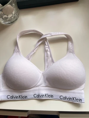 Vit sport-bh från Calvin Klein - Vit vadderad sport-bh från Calvin Klein med brottarrygg och bred resår med logga runt om. Tillverkad i mjukt och stretchigt material för extra komfort. Klassisk och stilren design som passar perfekt till träning eller som vardags-bh.