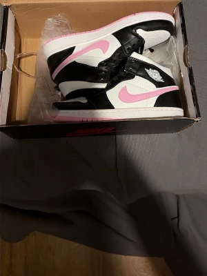 Nike Air Jordan 1 Mid svart/rosa - Snygga Nike Air Jordan 1 Mid sneakers i svart och vitt med rosa detaljer och sula. Klassisk siluett med läderöverdel, rosa swoosh och Wings-logga på sidan. Perfekta för dig som vill sticka ut med stilrena färgkombinationer och ikonisk design.