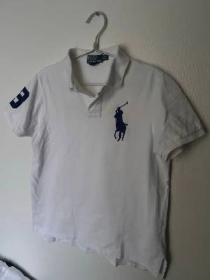 Vit pikétröja från Ralph Lauren - Klassisk vit pikétröja från Ralph Lauren med blå logga på bröstet och en stor blå siffra 3 på ena ärmen. Tröjan har krage, korta ärmar och knappar vid halsen. Perfekt till en avslappnad men ändå stilren look.