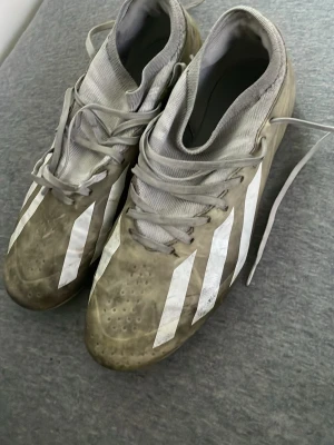  Adidas fotbollsskor med dobbar - Säljer ett par vita Adidas fotbollsskor med klassiska vita ränder på sidorna. Skorna har snörning, högre strumpkrage och slitstark yttersula med fasta dobbar för bra grepp på planen. Perfekta för dig som vill ha stilrena och funktionella skor till matchen.