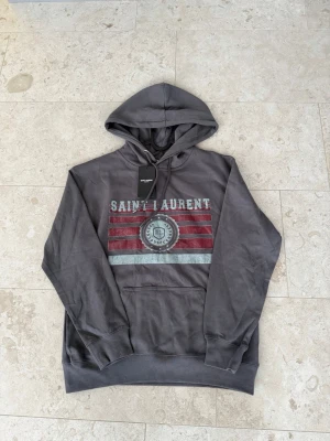 Grå Saint Laurent hoodie - Snygg grå hoodie från Saint Laurent med huva och dragsko. Framsidan har ett stort tryck med texten 'SAINT LAURENT' och röda samt vita ränder. (Kvitto finns)