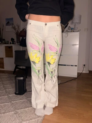 Vita bootcut jeans - Unika vintage vita jeans med målat blommotiv i gult, rosa och grönt på framsidan. Byxorna är bootcut och klassisk femficksmodell. Perfekta för dig som vill sticka ut med färgglada detaljer och konstnärlig vibe. de är gjorda i tunt jeans material som är perfekt inför sommaren. 