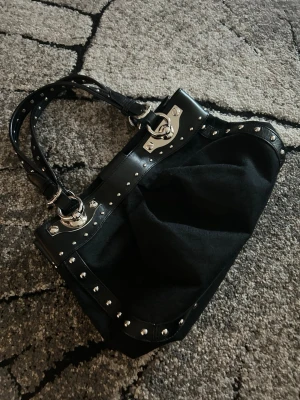 DKNY bag - Cool svart handväska med silvriga nitar och metallbeslag. Väskan har dubbla handtag i svart konstläder och detaljer i metall runt kanterna. Materialet är en mix av tyg och konstläder vilket ger en edgy look. Perfekt för dig som gillar en lite tuffare stil.