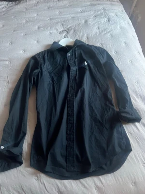 Svart skjorta från Polo Ralph Lauren - Klassisk svart skjorta från Polo Ralph Lauren i slim fit-modell. Skjortan har långa ärmar, knappar framtill och den ikoniska vita logobroderingen på bröstet. Perfekt för en stilren och enkel look.