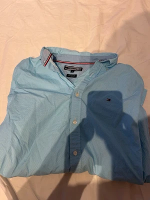 Ljusblå skjorta från Tommy Hilfiger - Snygg ljusblå skjorta från Tommy Hilfiger med klassisk button-down krage och diskret logga på bröstet. Skjortan har långa ärmar, vita knappar och är tillverkad i bomull. Perfekt för en clean och stilren look.