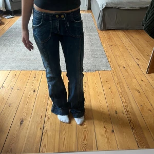 Mörkblå bootcut jeans med dubbla knappar - Säljer ett par mörkblå bootcut jeans med dubbla knappar i midjan och dekorativa sömmar. Jeansen har låg midja, stora fickor fram och bak samt ljusa slitningar för en cool look. Perfekta för dig som gillar retrostil och vill ha ett par unika jeans. Står ingen storlek men jag brukar ha storlek S