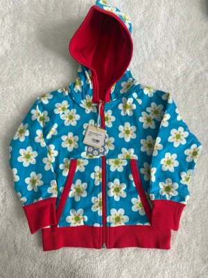 Blommig hoodie från Maxomorra - Färgglad hoodie  från Maxomorra med blå botten och stora vita blommor över hela plagget. Röd huva, muddar och fickor ger en snygg kontrast. Dragkedja framtill och mjukt bomullsmaterial gör den både bekväm och stilren. Perfekt för dig som gillar lekfulla mönster! Man kan använda båda sidor . 