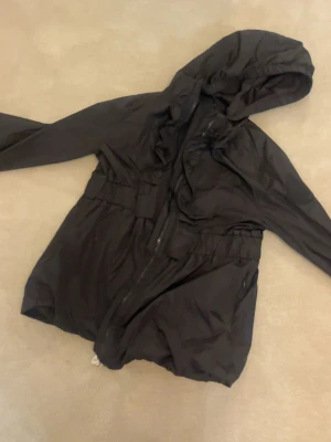 Svart vindjacka från Moncler - Snygg svart vindjacka från Moncler med huva och dragkedja framtill. Jackan har elastisk midja för en schysst passform och praktiska fickor med dragkedja. Tillverkad i lätt, vindtätt material som passar perfekt för blåsiga dagar.