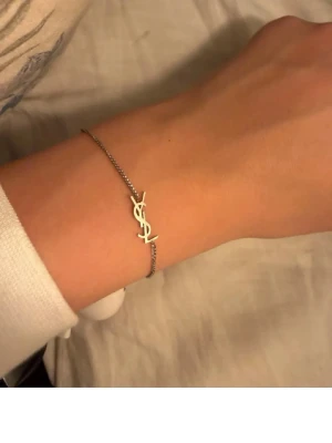 Silverfärgat YSL armband - Snyggt och stilrent armband från Yves Saint Laurent i silverfärg. Tunt kedjearmband med den ikoniska YSL-logotypen i mitten. Perfekt accessoar för dig som gillar minimalistisk och lyxig design.