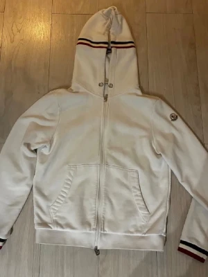 Vit hoodie från Moncler med dragkedja - Snygg vit hoodie från Moncler med hel dragkedja, huva och fickor framtill. Huva och ärmslut har röd-svart randiga detaljer. Moncler-logga på vänster ärm. Perfekt för en stilren look
