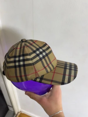 En äkta beige Burberry keps med rutmönster - Säljer en klassisk beige keps från Burberry med det ikoniska rutmönstret i svart, vitt och rött. Kepsen har böjd skärm och justerbar rem bak. Perfekt accessoar för dig som vill ha en tidlös och trendig look.