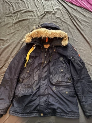 Mörkblå Parajumpers parkas med pälskant - Säljer en mörkblå parkas från Parajumpers med avtagbar huva och pälskant. Jackan har flera praktiska fickor, dragkedjor och den ikoniska gula remmen vid kragen. Perfekt för kalla dagar och riktigt snygg streetstil. Materialet är slitstarkt och vattenavvisande. Jackan är använd men fortfarande väldigt bra skick då dessa jackor är väldigt hållbara. Köpt ny för närmare 10 tusen kronor så priset känns rimligt med tanke på det bra skicket men kan fortfarande diskuteras. Skriv gärna vid frågor!💙