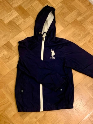 Marinblå vindjacka från U.S. Polo Assn. - Snygg marinblå vindjacka från U.S. Polo Assn. med vit dragkedja och logga på bröstet. Jackan har huva, meshfoder och två fickor med knappstängning. Perfekt för blåsiga dagar och enkel att matcha till olika outfits. Köpt i USA. (Storlek S i barnstorlek)