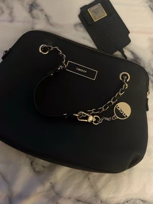 Svart handväska från DKNY med kedjedetalj (Donna karen New York)  - Snygg svart handväska från DKNY med guldfärgade metalldetaljer och kedjehandtag. Väskan har en stilren rektangulär form, dragkedja upptill och DKNY-logga framtill. Tillverkad i slätt konstläder och kommer med matchande nyckelring. Väldigt bra skick och fin 