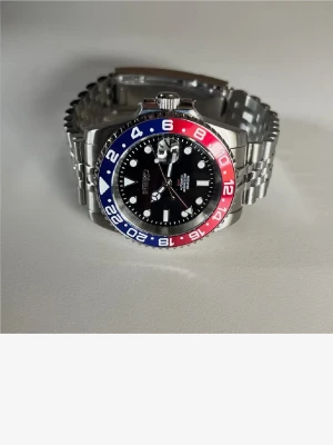 Seiko klocka med Pepsi-bezel - Stilren Seiko klocka med klassisk Pepsi-bezel i blått och rött. Klockan har svart urtavla, tydliga index och datumvisning med förstoringsglas. Länkarmband i silverfärgat stål ger en sportig och tidlös look.