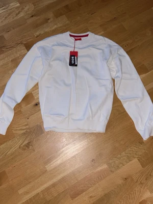 Vit sweatshirt från BOSS - Säljer en stilren vit sweatshirt från BOSS med rund halsringning och ribbade muddar vid ärmslut och nederkant. Klassisk passform och diskret design, perfekt för dig som gillar minimalistisk stil. Materialet känns mjukt och bekvämt mot huden.