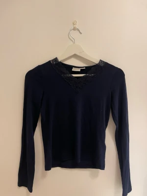 Mörkblå topp med spetsdetalj - Mörkblå långärmad topp med snygg spetsdetalj i v-ringning. Toppens passform är normal och materialet känns mjukt och bekvämt. Perfekt för dig som gillar stilrena plagg med en twist.