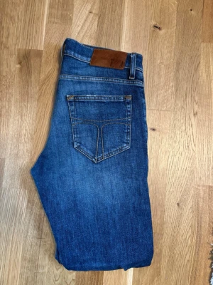 Tigerofsweden pistolero jeans - Snygga blå jeans med klassisk femficksdesign och raka ben. Jeansen har en brun läderpatch i midjan och kontrastsömmar. Perfekta för en avslappnad och trendig look. Materialet är denim i bomull.
