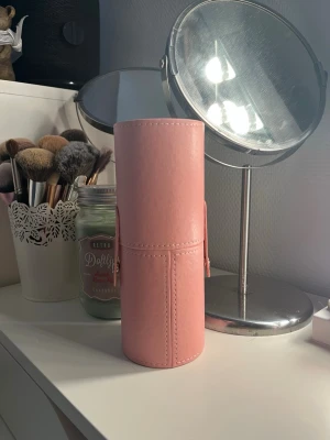 Rosa cylinderformad borsthållare - Praktisk och stilren cylinderformad borsthållare i rosa konstläder. Perfekt för att förvara och skydda sminkborstar eller andra skönhetsverktyg. Hållaren har tryckknappar på sidorna och kan delas i två delar för enkel åtkomst.