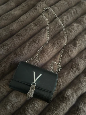 Svart axelväska från Valentino - Snygg svart axelväska från Valentino med silverfärgad kedjerem och dekorativt V-spänne framtill. Väskan är köpt för 900, den har lite skavanker som ni kan se på bild två🫶🏼