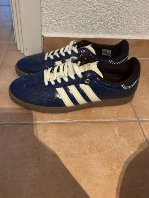 Adidas Wales Bonner  - Snygga blå sneakers från Adidas med vita ränder på sidorna och vita skosnören. Skorna har ett unikt krokodil-liknande mönster i lädret och bruna detaljer på insidan samt gummisula. Perfekta för dig som vill sticka ut med klassisk streetstyle.