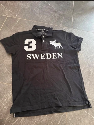 Svart pikétröja med tryck SWEDEN - Svart pikétröja från Swedemount med vit text 'SWEDEN', en stor siffra 3 och en älg på bröstet. Klassisk krage med knappar och korta ärmar. Perfekt för dig som vill ha en sportig och stilren look.