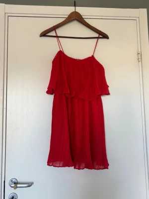Röd plisserad kort klänning - Snygg röd klänning med tunna axelband och plisserat tyg. Klänningen är kort och har en volang upptill som ger en extra touch. Har blivit lite tråddragningar i tyget som visas 