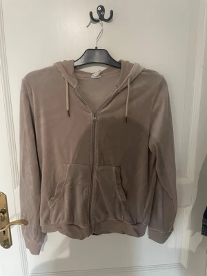 Beige hoodie från Female Passion - Mysig beige hoodie från Female Passion med dragkedja framtill, två fickor och justerbar huva med snören. Tillverkad i ett mjukt, sammetsliknande material som känns skönt mot huden. Perfekt för en avslappnad och chill stil.