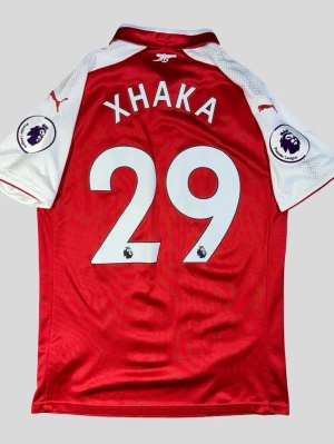 Äkta 2017-18 Arsenal XHAKA 29 home - M  - Äkta 2017-18 Arsenal XHAKA 29 home - M   Puma Arsenal matchtröja med spelarnamn Xhaka och nummer 29. Tröjan är röd med vita ärmar och har Arsenal-klubbmärke, Puma-loggor och Premier League-märken på ärmarna. Sponsorlogga på bröstet. Kortärmad modell i lätt funktionsmaterial, lämplig för fotboll och annan träning eller som supportertröja.  Kan skickas mot frakt kostnad OBS FINNS UTE PÅ FLER SIDOR
