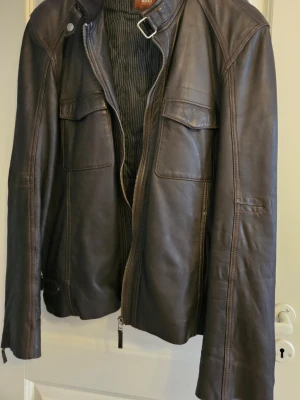 Mörkbrun skinnjacka från Saki - Snygg mörkbrun skinnjacka från Saki med två bröstfickor med lock, ståkrage med spänne och dragkedja framtill. Jackan har raka ärmar och markerade sömmar för en cool look. Perfekt för dig som vill ha en klassisk men trendig stil.