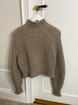 Beige chunky stickad polotröja - Säljer en beige chunky stickad polotröja med grov ribbstickning. Tröjan har hög krage, långa ärmar och avslappnad passform. Perfekt för kalla dagar och enkel att styla med jeans eller kjol.