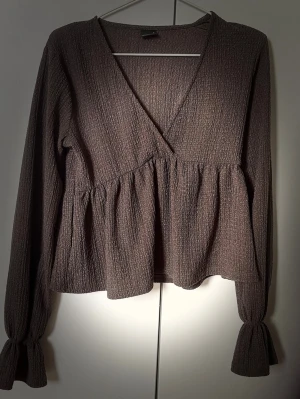 Brun blus - Säljer en brun blus med v-ringning och volangdetaljer. Blusen har långärmade ärmar med resår vid ärmsluten som ger en ballongeffekt. Perfekt att styla med jeans eller kjol.
