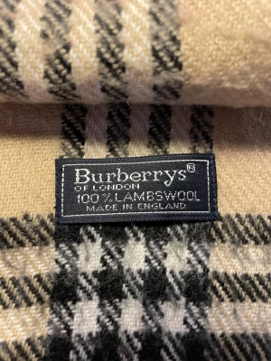 Beige rutig halsduk från Burberrys - Klassisk beige halsduk från Burberrys i 100% lammull med det ikoniska rutmönstret i svart, vitt och rött. Halsduken har fransar i ändarna och är tillverkad i England. Perfekt accessoar för att lyfta din outfit.