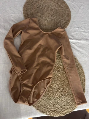 Brun långärmad konståknings body - Snygg beige body med långa ärmar och rund halsringning. Tillverkad i ett stretchigt och mjukt material som sitter skönt mot kroppen. Storlek 140 