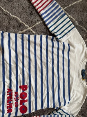 Randig långärmad t-shirt Polo Atelier - Snygg långärmad t-shirt från Polo Ralph Lauren Atelier med blå och vita ränder samt färgglada ränder på ärmarna. Stor röd logga framtill. Tillverkad i mjuk bomull, perfekt för en avslappnad stil.