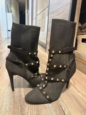Svarta stövletter med nitar Bella Star - Snygga svarta stövletter från Bella Star med höga smala klackar och stickad textil. Dekorerade med remmar och guldfärgade nitar för en edgy look. Stövletterna har spetsig tå och åtsittande skaft i stretchigt material. Använts några gånger men inte något tecken på att de är skadade