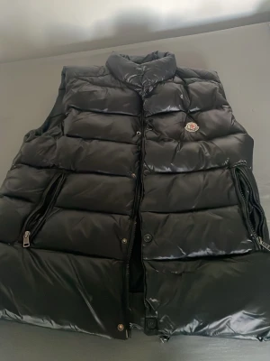 Svart dunväst från Moncler - Snygg svart dunväst från Moncler med glansig finish, hög krage och två dragkedjefickor. Klassisk Moncler-logga på bröstet och robusta dragkedjor. Perfekt för lager-på-lager och streetstyle.