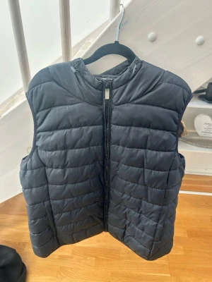 Svart dunväst med huva - Snygg svart dunväst med huva och dragkedja framtill. Västen har quiltad design med horisontella sömmar och är perfekt för lager-på-lager. Tillverkad i ett lätt och värmande material som håller dig varm under kyliga dagar.