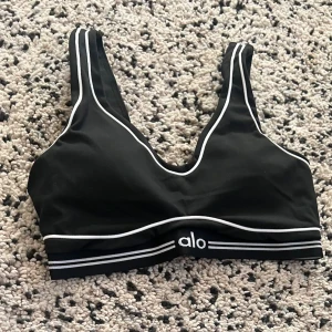 Svart sport-bh från Alo med vita detaljer - Sportig svart bh från Alo med vita kontrastsömmar och bred resår med logga. Mjuk och bekväm modell utan byglar, perfekt för träning eller chill. Snygg V-ringning och breda axelband för extra stöd.