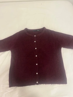 Vinröd kofta med pärlknappar - Mysig vinröd kofta med rund halsringning och pärlknappar framtill. Koftan är stickad i ett mjukt material som känns skönt mot huden. 