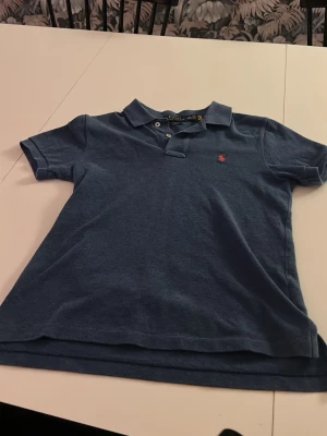 Blå pikétröja från Ralph Lauren 10-12 - Snygg blå pikétröja från Ralph Lauren med klassisk krage och två knappar. Tröjan har korta ärmar och en liten röd broderad logga på bröstet. Perfekt för en avslappnad och stilren look.