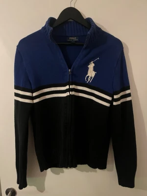 Blå och svart ziptröja från Polo Ralph Lauren - Snygg ziptröja i blått och svart från Polo Ralph Lauren med vita ränder över bröstet och stor broderad logga på vänster sida. Tröjan har hög krage och ribbade muddar. Perfekt för en sportig och stilren look.