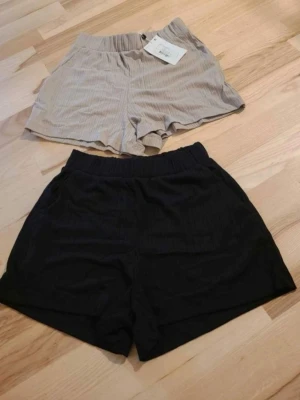 Två par randiga shorts i svart och beige - Två par shorts i mjukt tyg, ett par i svart och ett i beige. Båda har diskreta ränder och elastisk midja för skön passform. Perfekta för varma dagar och enkel stil. Säljs tillsammans.
