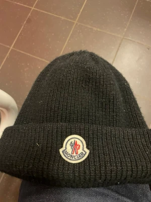 Svart Moncler mössa med logga - Säljer en svart ribbstickad mössa från Moncler med uppvikt kant och den klassiska Moncler-loggan framtill. Mössan är i mjukt material och har en clean, stilren look som passar perfekt till höst och vinter.