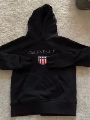Svart hoodie från GANT med logga - Säljer en svart hoodie från GANT med stor huva och känguruficka. På bröstet finns GANT-loggan och ett broderat sköldmärke i rött, vitt och blått. Hoodien har ribbade muddar och är gjord i mjukt bomullsmaterial. Perfekt för en chill och stilren look.